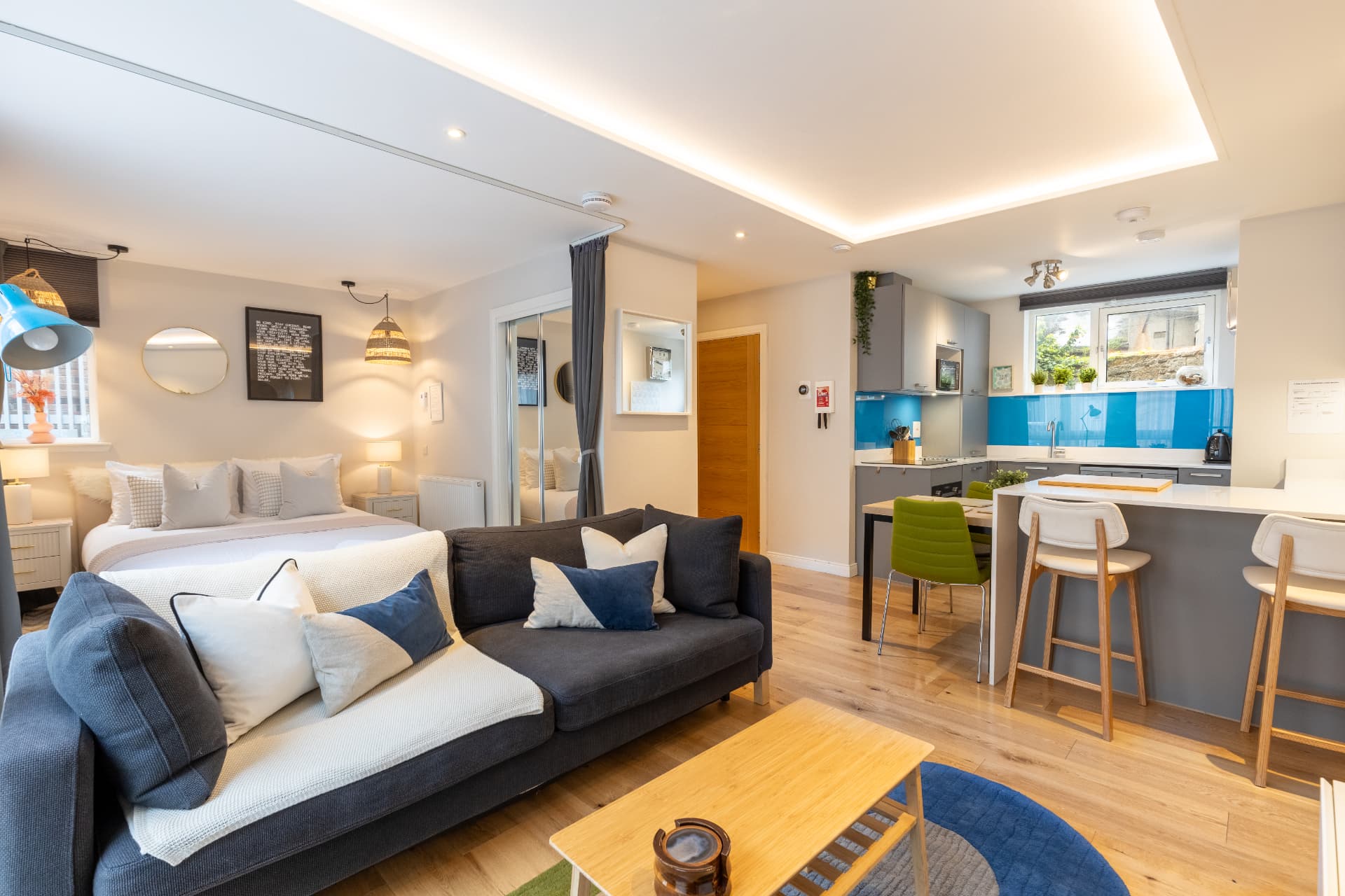 Open-plan interior at Heavenly Pad, with a grey sofa, sleeping area and kitchen with a blue splashback.